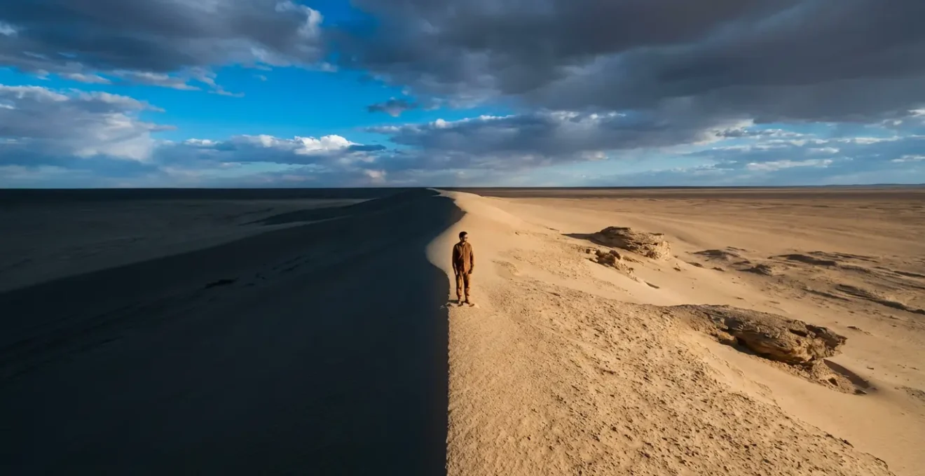 Wide landscape showing solitary figure standing between dramatic light and shadow zones in minimalist desert