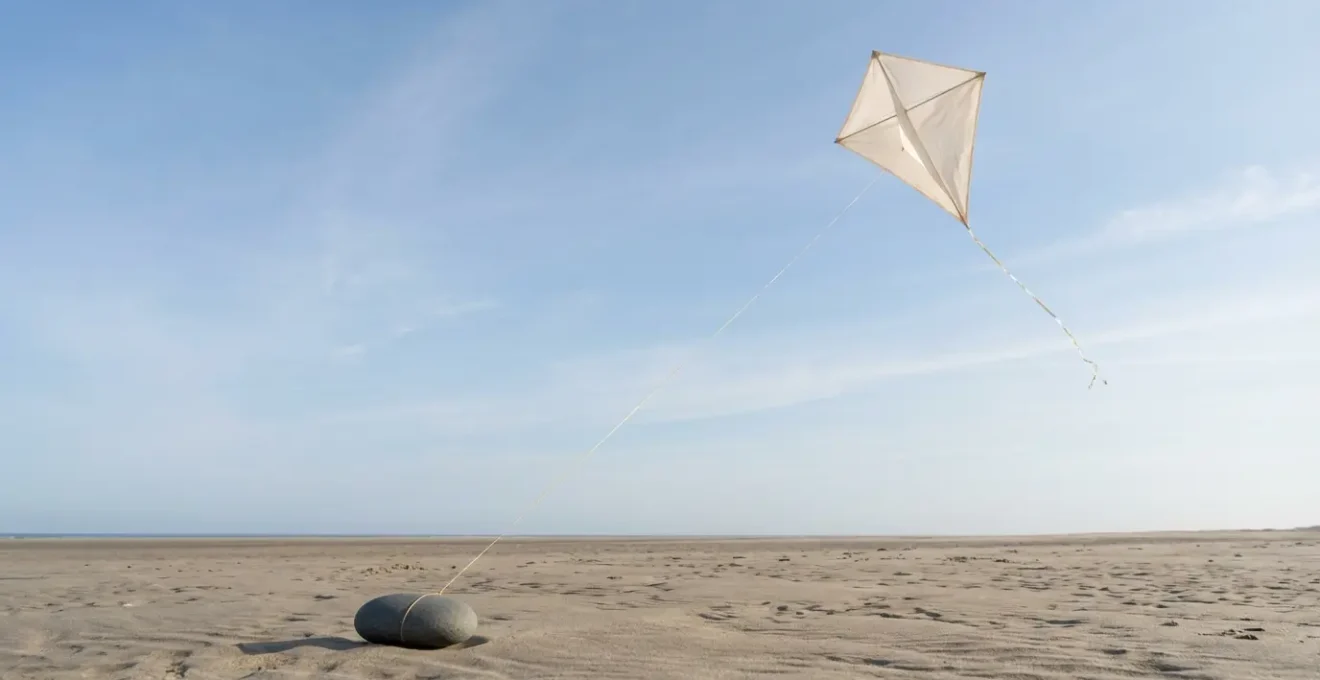 A delicate kite tethered by a golden thread to a smooth stone anchor on a sandy beach, representing the balance of freedom and stability.